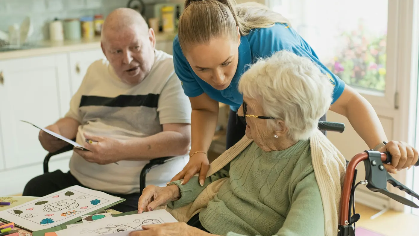 A caregiver helping two older adults with a coloring activity at a table